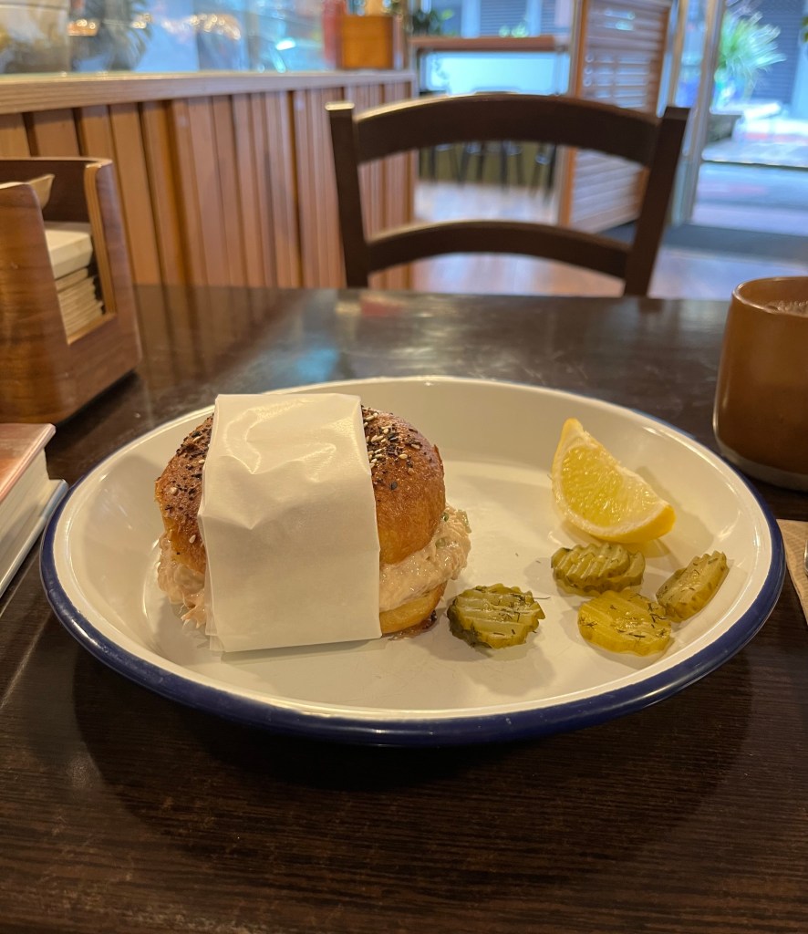 A plate featuring a tuna salad bagel wrapped in paper, accompanied by a slice of lemon and pickles, set on a wooden table in a cafe.