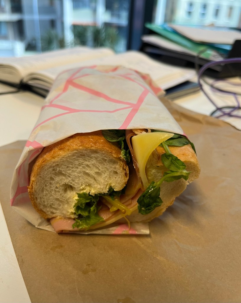 A close-up view of a sandwich filled with ham, cheese, and green leaves, wrapped in pink and white paper, placed on a brown surface with blurred office items in the background.